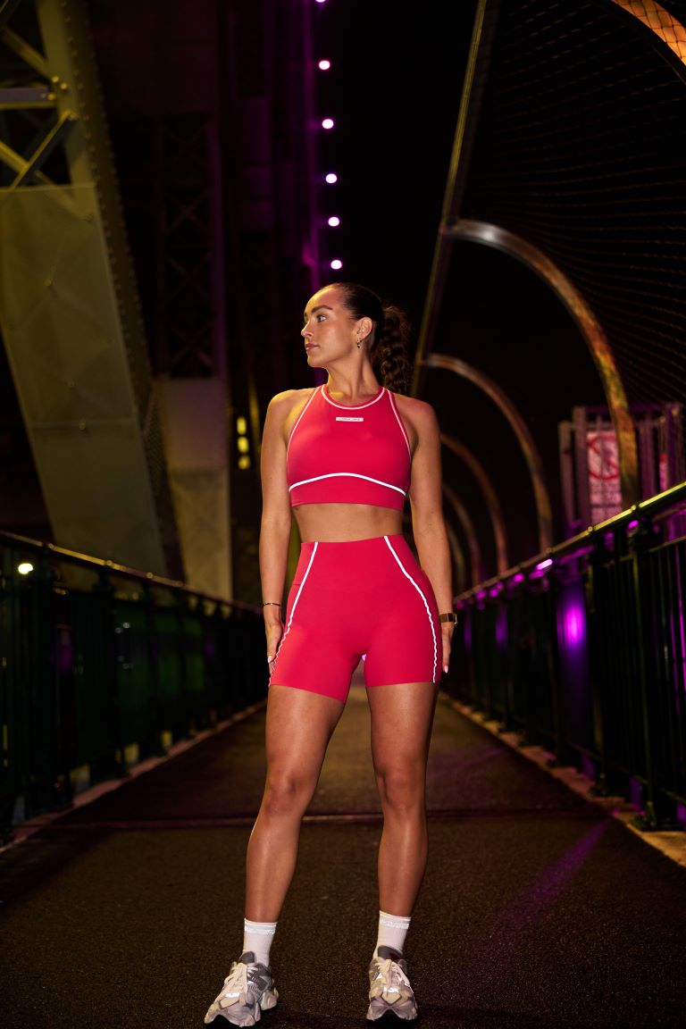 Caity Viant standing on the Brisbane Story Bridge pedestrian walkway wearing matching Lorna Jane Night Runner Sports Bra and 16cm Bike Shorts in Neon Raspberry from the new Reflective Run Collection