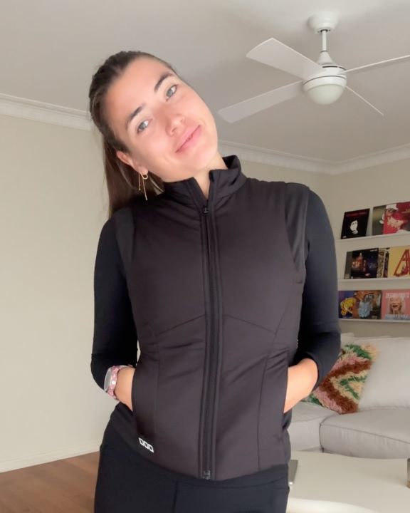 Selfie of Eliza Boyd in her living room cocking her head to one side wearing a black vest, long sleeve top and leggings