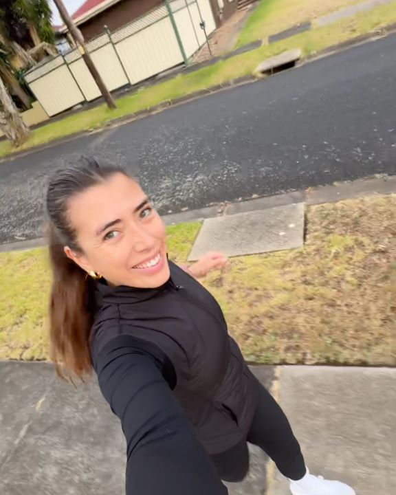 Selfie of Eliza Boyd running on a street sidewalk smiling at the camera wearing a black vest, long sleeve top, leggings and white sneakers